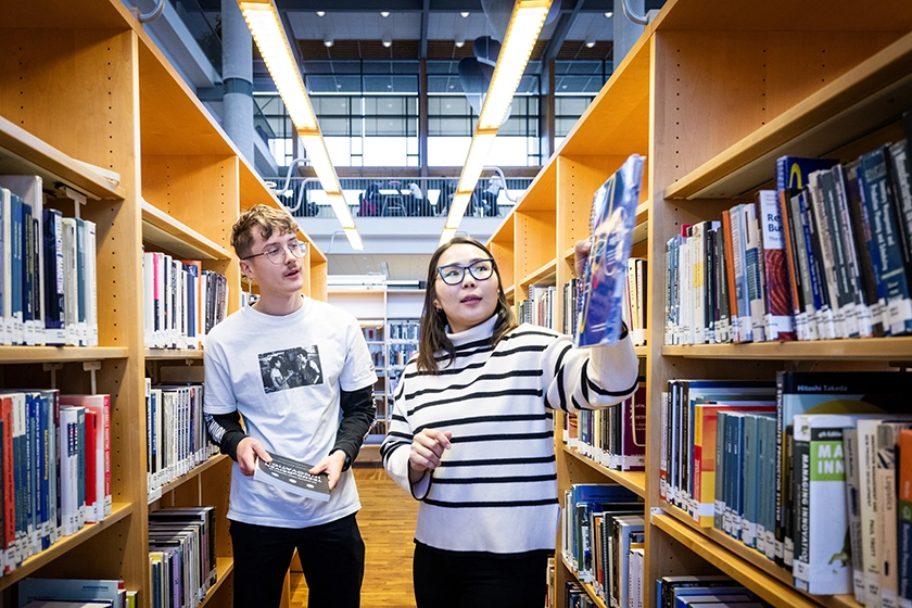 Två studenter i biblioteket som står mellan bokhyllor och håller i böcker.